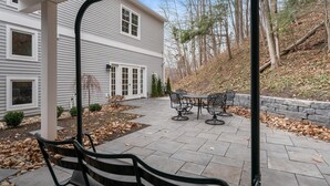 Une terrasse avec des meubles d’extérieur, un chemin en pierre et une maison aux murs recouverts de bardage blanc.