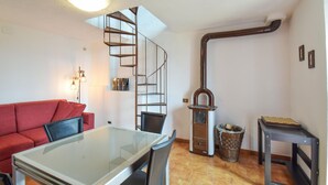 Un salon chaleureux avec un canapé rouge, une table à manger avec un plateau en verre, un escalier en colimaçon, un poêle à bois et un jeu d’échecs.