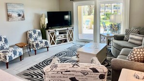 A living room with a TV, patterned furniture, a ceiling fan, and a view of the outdoors.
