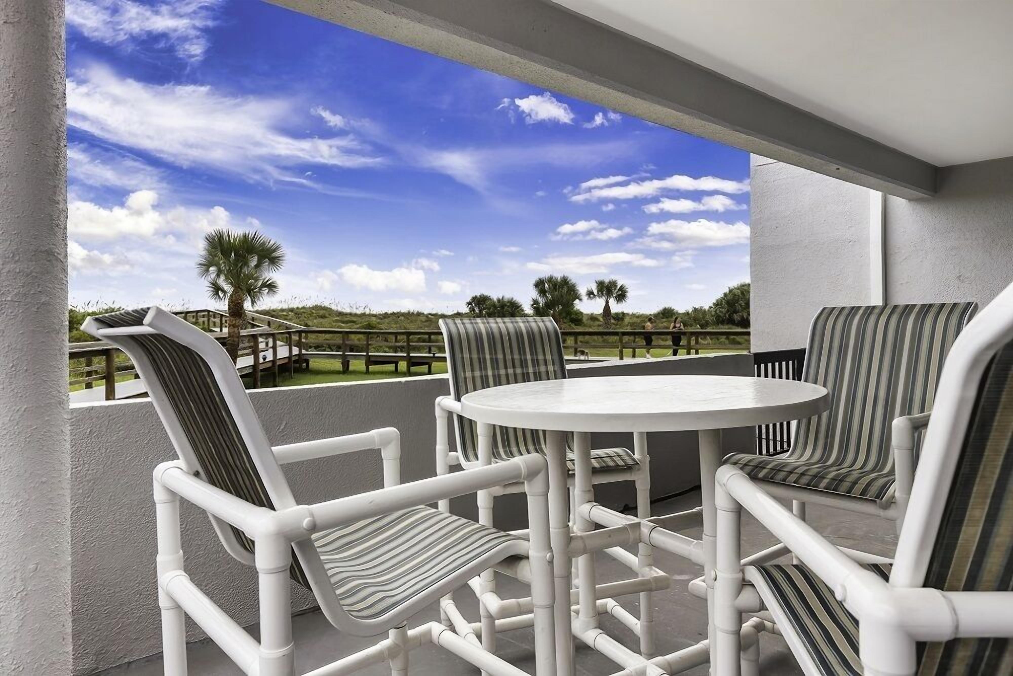 A patio area with white chairs and a round table, overlooking a grassy landscape with palm trees.