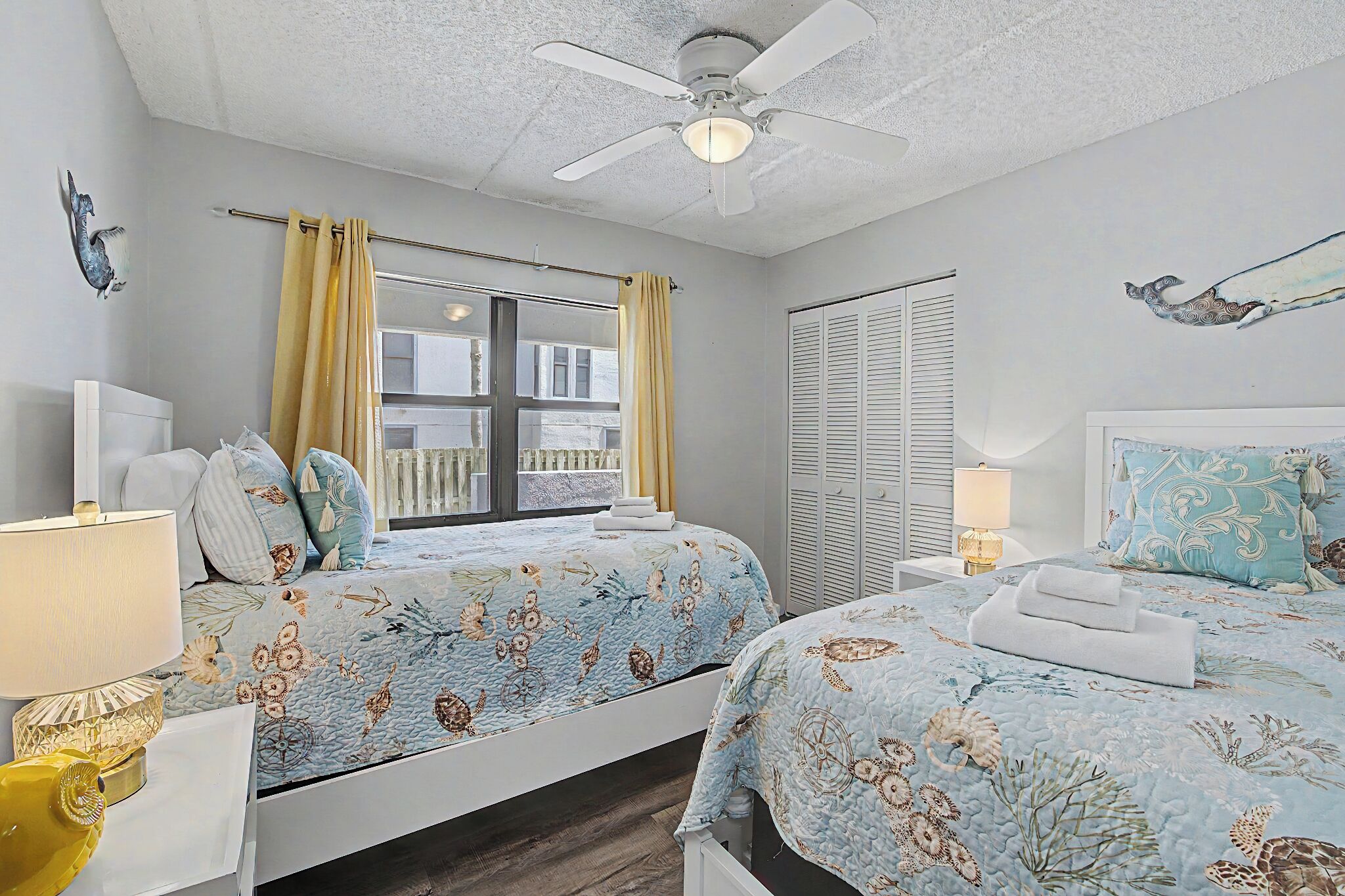Une chambre avec deux lits, un ventilateur de plafond, une fenêtre avec des rideaux, un radiateur et un luminaire fixé au mur.