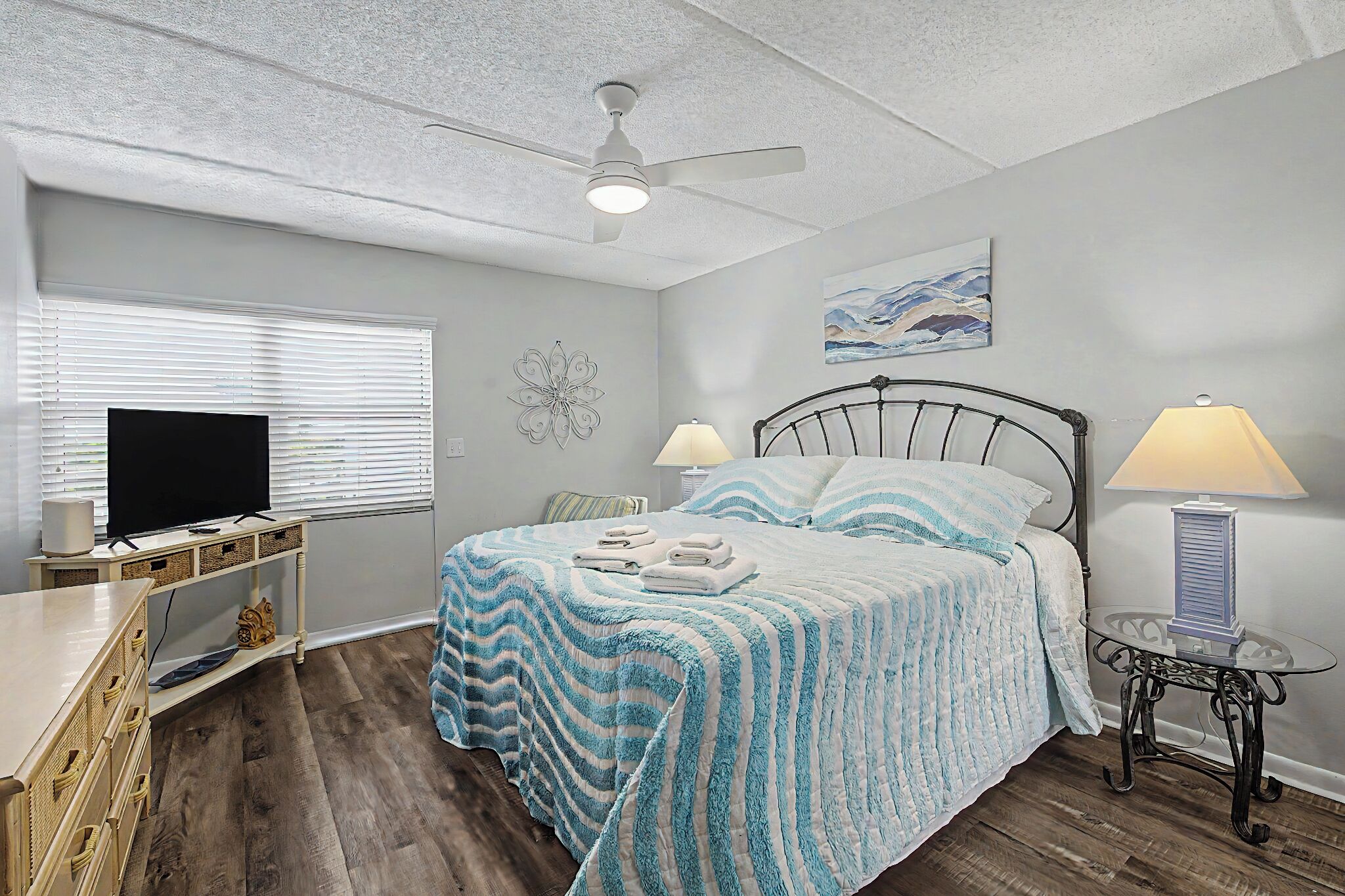 A bedroom with a bed, a television, a ceiling fan, a bedside table with a lamp, and a wall art piece.