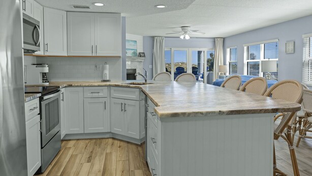 A modern kitchen with white cabinets, a large island, and a dining area with wicker chairs. - Ground Floor Oceanfront Condo With Beach Access At Your Back Door! (St. Augustine)