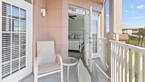 Un balcone con una sedia e un tavolo bianchi, vista su una camera da letto con un letto e una ventola a soffitto, e un cielo sereno.