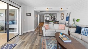 A spacious living area with a sliding glass door leading to a balcony, a sofa set, a coffee table, and a kitchen area in the background. - Stunning views from this top floor three bedroom condo! (Saint Augustine)