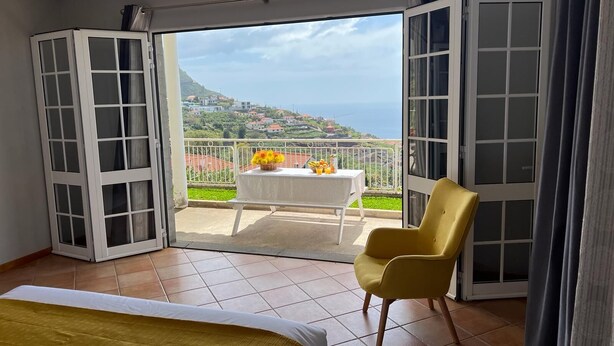 A bedroom with a bed, a yellow chair, a table with fruits and flowers, and a balcony with a view of houses and the sea. - Spring House AL (Arco da Calheta)