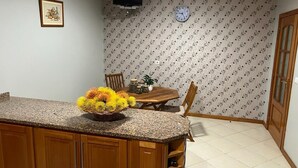 A kitchen with a granite countertop, wooden cabinets, a dining table with chairs, a clock on the wall, and patterned wallpaper. - Spring House AL (Arco da Calheta)