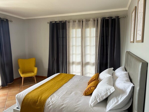 Un dormitorio ordenado con una cama, un sillón amarillo y una ventana con cortinas.