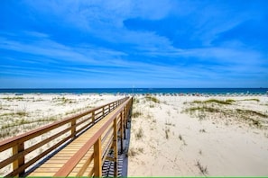 Une promenade en bois qui mène à une plage de sable, bordée de dunes, sous un ciel bleu et dégagé.