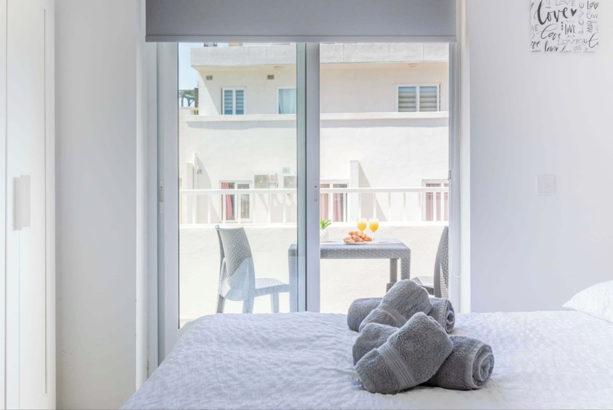 Habitación de hotel con cama, toallas, balcón con mesa y sillas, y vistas a edificios.