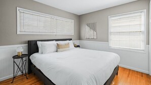 A bedroom with a large bed, two windows with blinds, a wall-mounted light fixture, and a small table with a lamp. - South Park Quiet 2bd Close to Downtown + AC (San Diego)