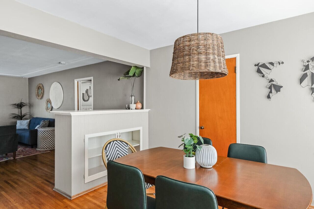 Uma sala de jantar com uma mesa de madeira, cadeiras verdes, uma luminária pendente de vime e obras de arte na parede.