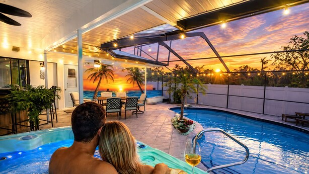A couple in a hot tub with a sunset view, a pool, and outdoor seating. - Heated Pool & Hot Tub | Game Room | Near AMI (Bradenton)