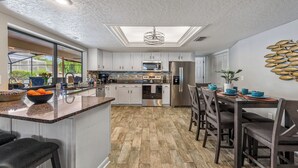 A modern kitchen with a central island, stainless steel appliances, and a dining area with a view of the outdoors. - Heated Pool & Hot Tub | Game Room | Near AMI (Bradenton)