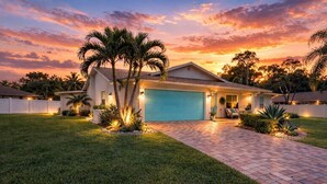 A single-story house with a blue garage door, surrounded by a white fence, palm trees, and a well-maintained lawn. - Heated Pool & Hot Tub | Game Room | Near AMI (Bradenton)