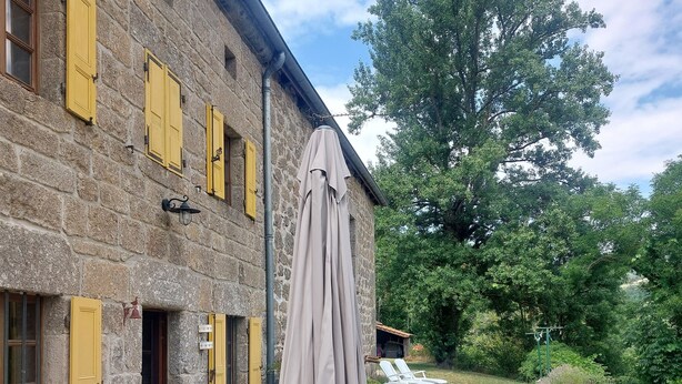 Un edificio de piedra con contraventanas amarillas, mobiliario exterior con una mesa y sillas, y un área cubierta con una sombrilla.