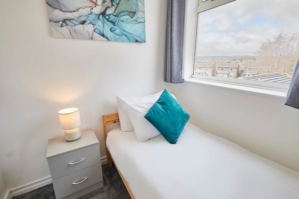 A bedroom with a bed, a nightstand with a lamp, a window with a view of buildings, and a marble artwork on the wall. - Host & Stay | Bartlemere House (Barnard Castle)
