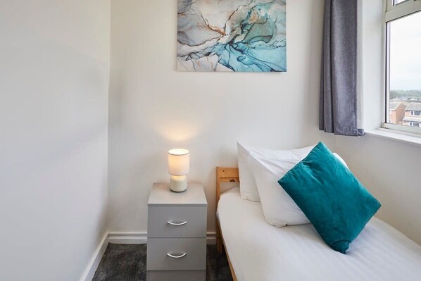 A bedroom with a bed, a nightstand with a lamp, a drawer, and a wall art piece.