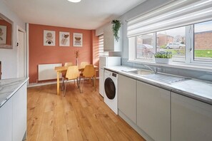 A modern kitchen with a built-in washing machine, a dining area with wooden chairs, and a view of the outside through large windows.
