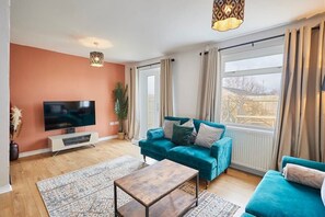 Ett vardagsrum med en turkos soffa, ett soffbord i trä, en platt-TV på ett vitt TV-bord och ett stort fönster med gardiner.