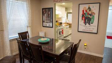 Un coin repas avec une table, des chaises et une fenêtre avec des stores. Un poster encadré est accroché au mur et une petite lampe de table est posée sur le sol.