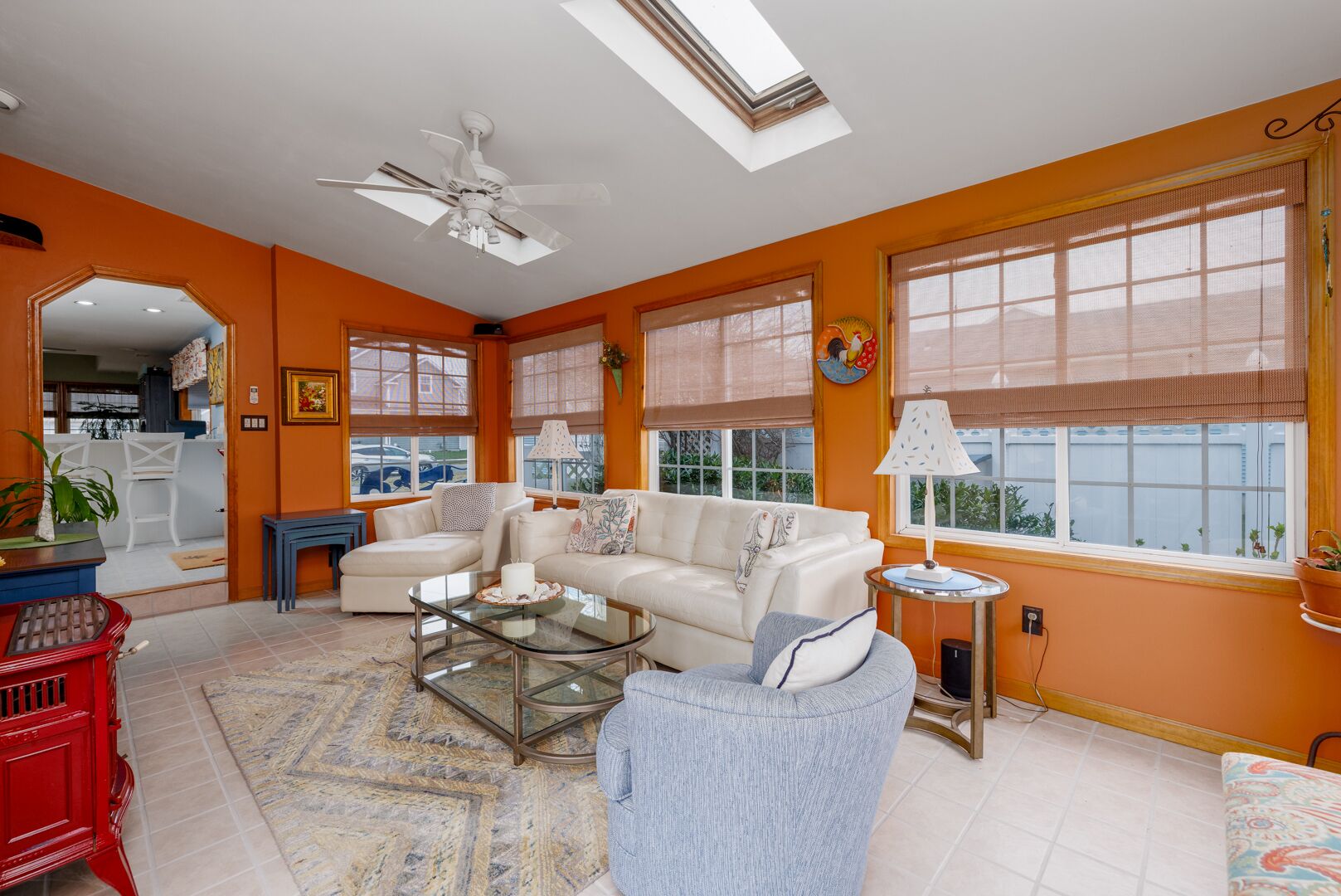 A spacious living room with a large sofa, a glass coffee table, and a ceiling fan. There are large windows with blinds, a red cabinet, and a small side table with a lamp.