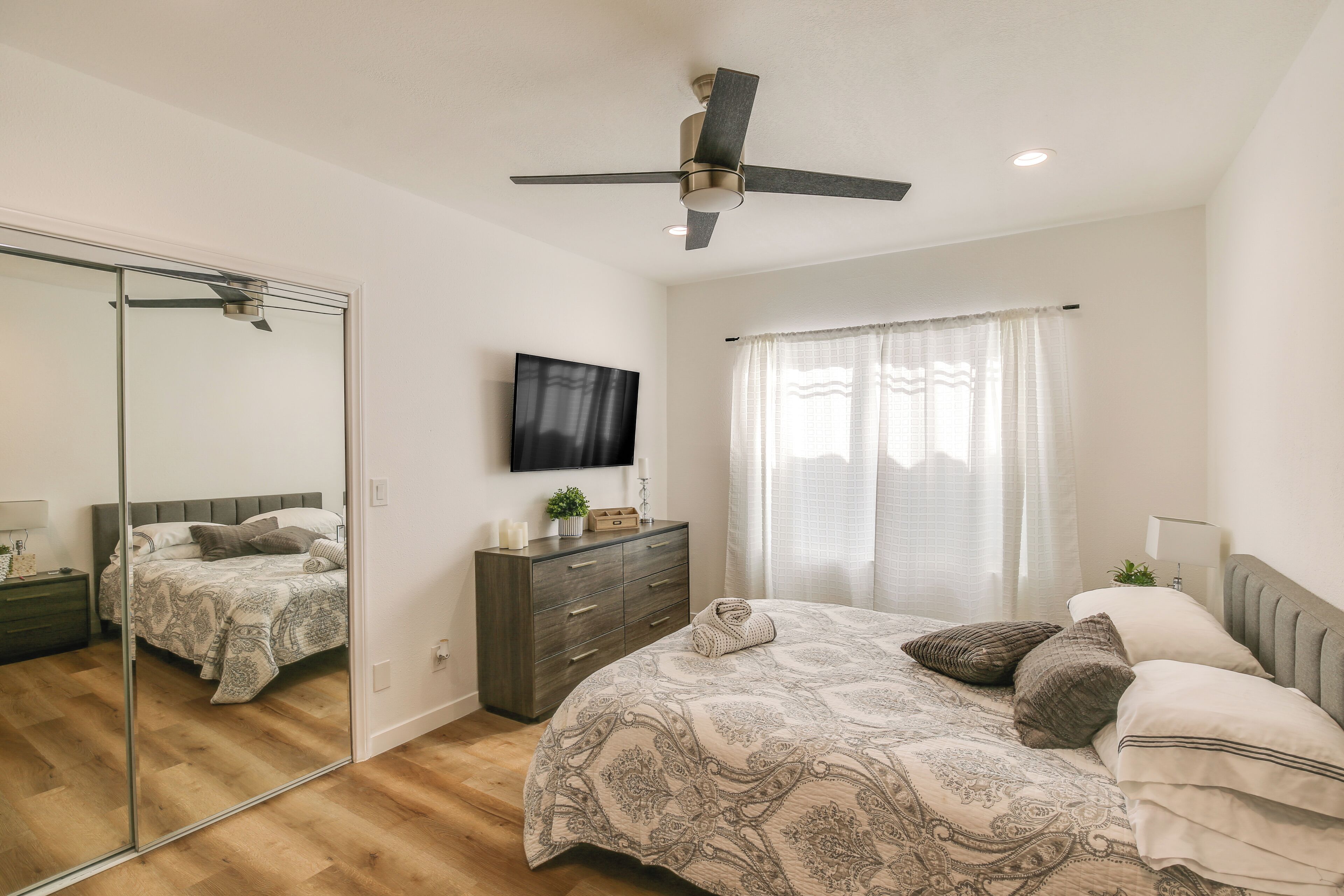 A bedroom with a bed, a dresser, a mirror, a ceiling fan, and a TV mounted on the wall.