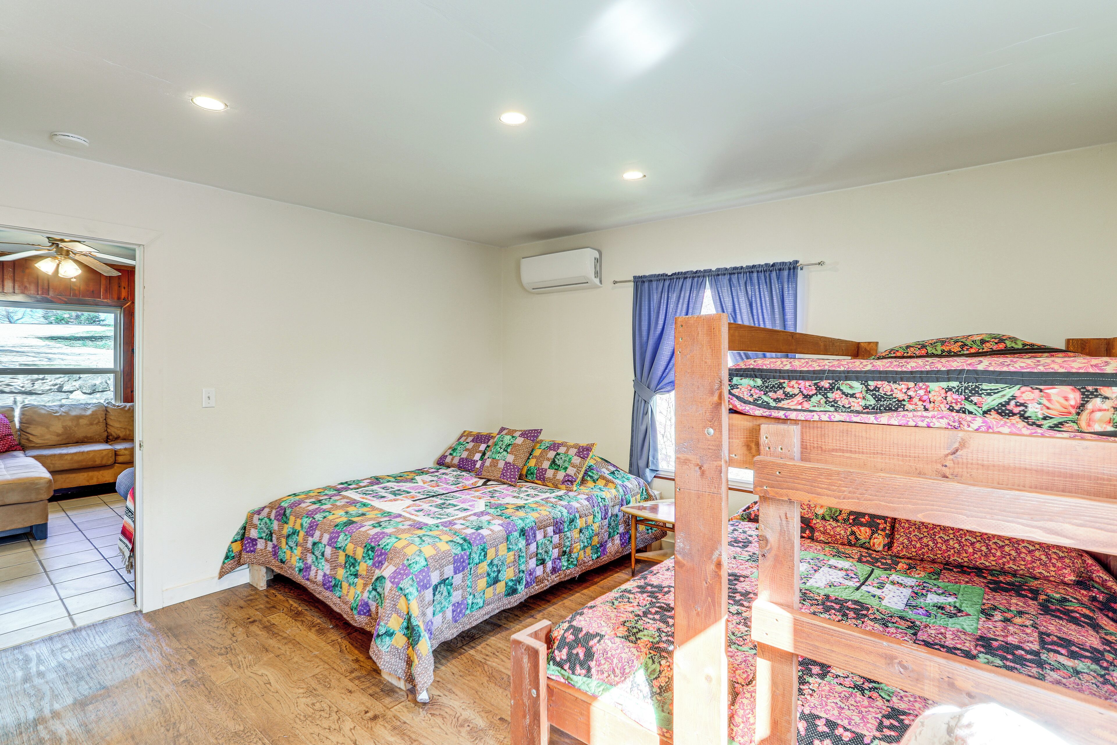 A bedroom with a wooden bunk bed, a single bed with a colorful quilt, a ceiling fan, and an air conditioning unit.