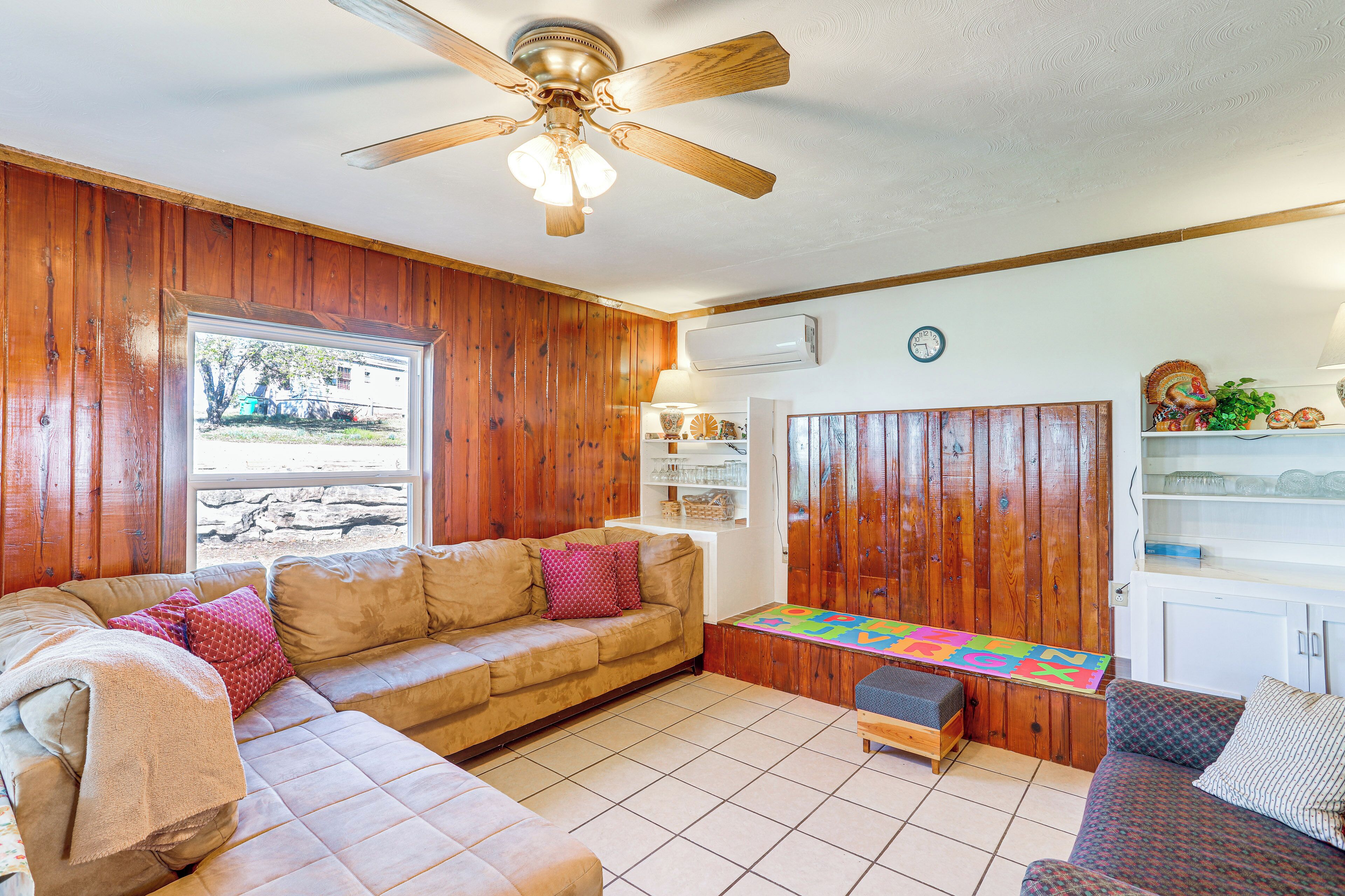 A cozy living room with wooden walls, a ceiling fan, a large window, a sofa, a small table, and a colorful play mat.