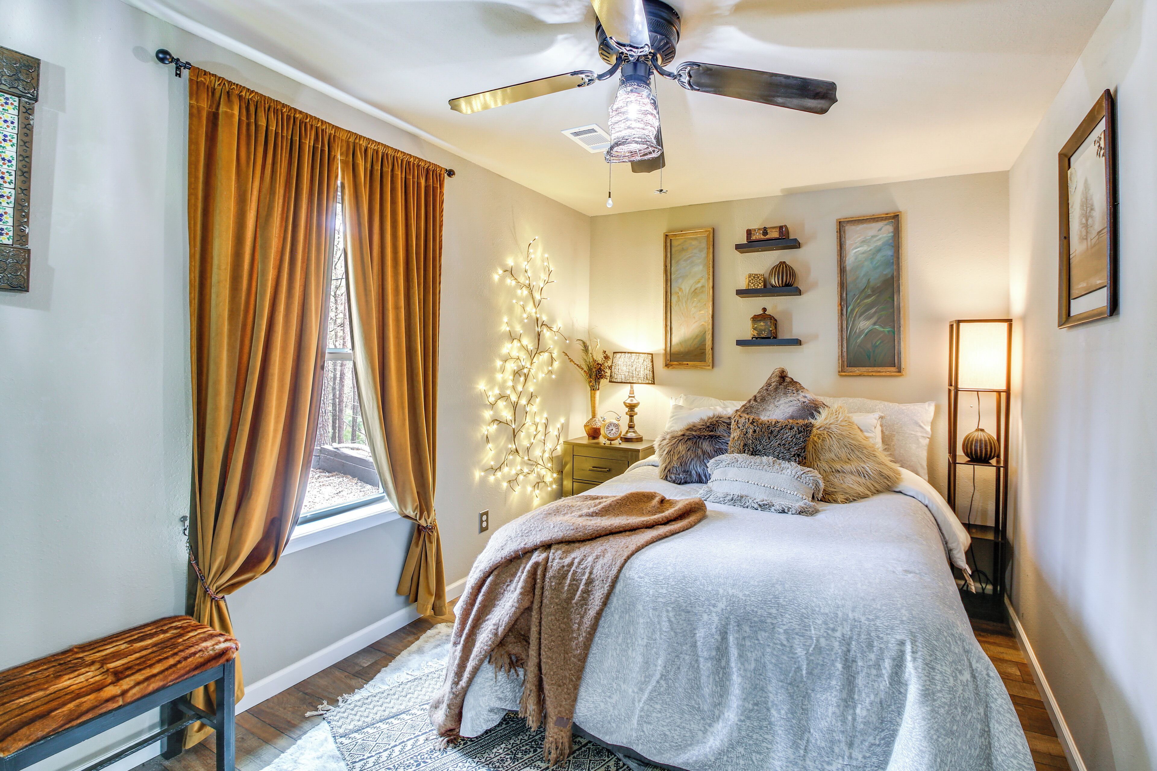 Un dormitorio con cama, ventilador de techo, un banco, una ventana con cortinas y estanterías.