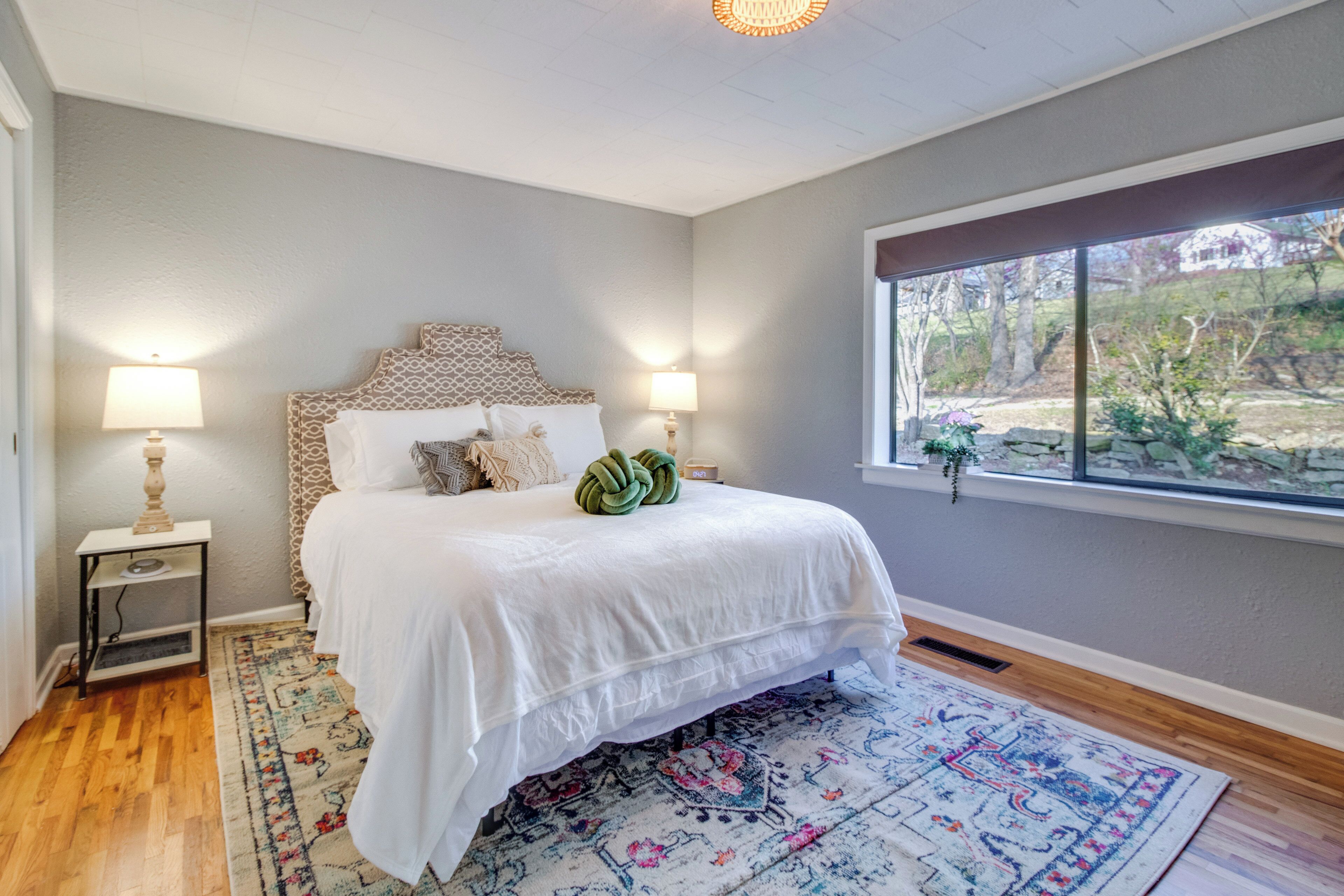 A bedroom with a large bed, two bedside lamps, a patterned rug, and a window with a view of greenery.