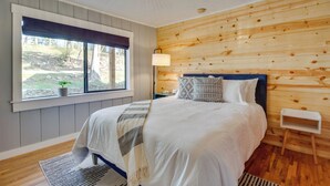 Une chambre avec un mur lambrissé en bois, un grand lit recouvert d’une couette blanche, une table de chevet, une lampe et une fenêtre donnant sur l’extérieur.
