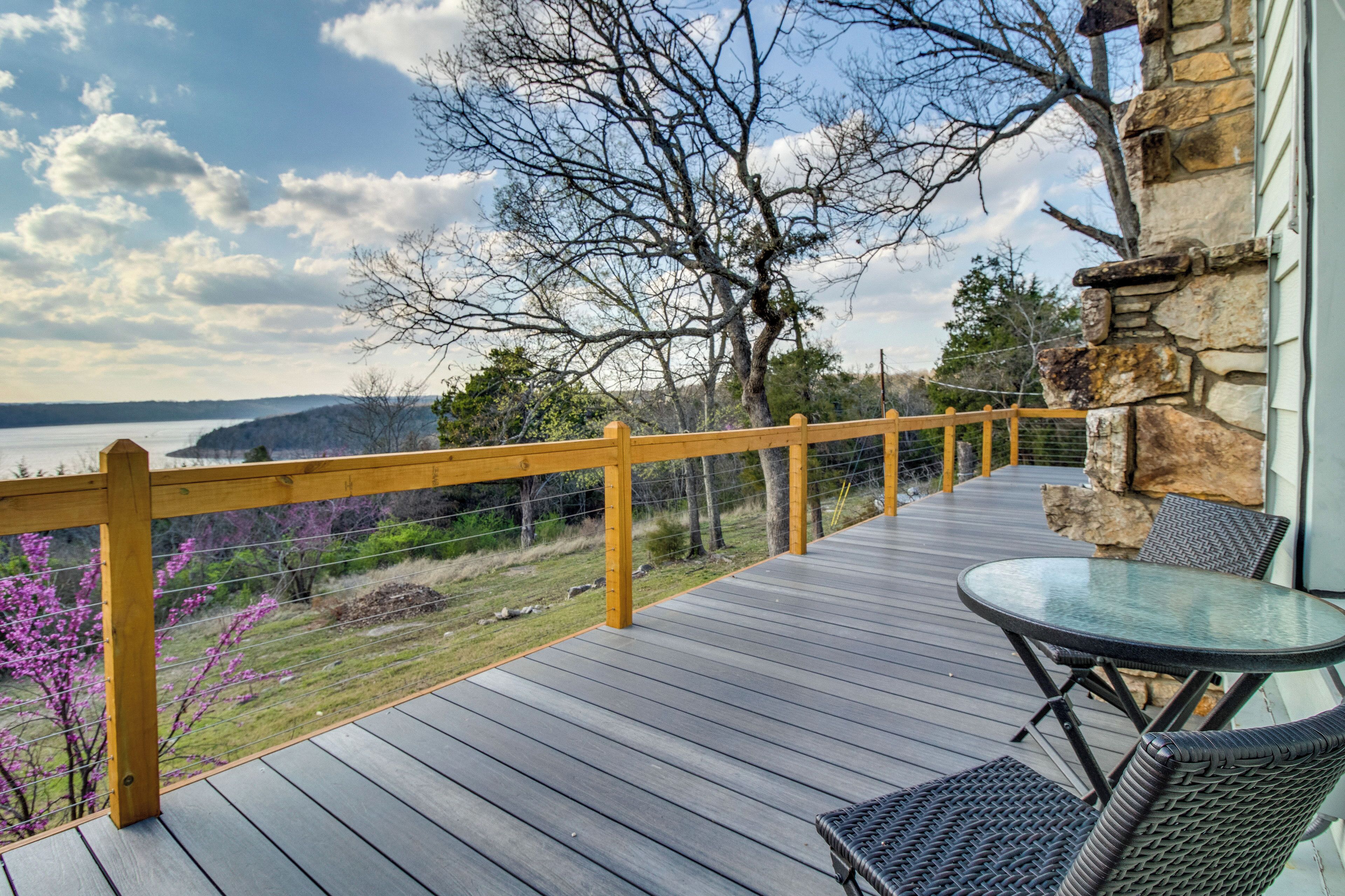 A wooden deck with a glass table and chairs, overlooking a body of water and hills.