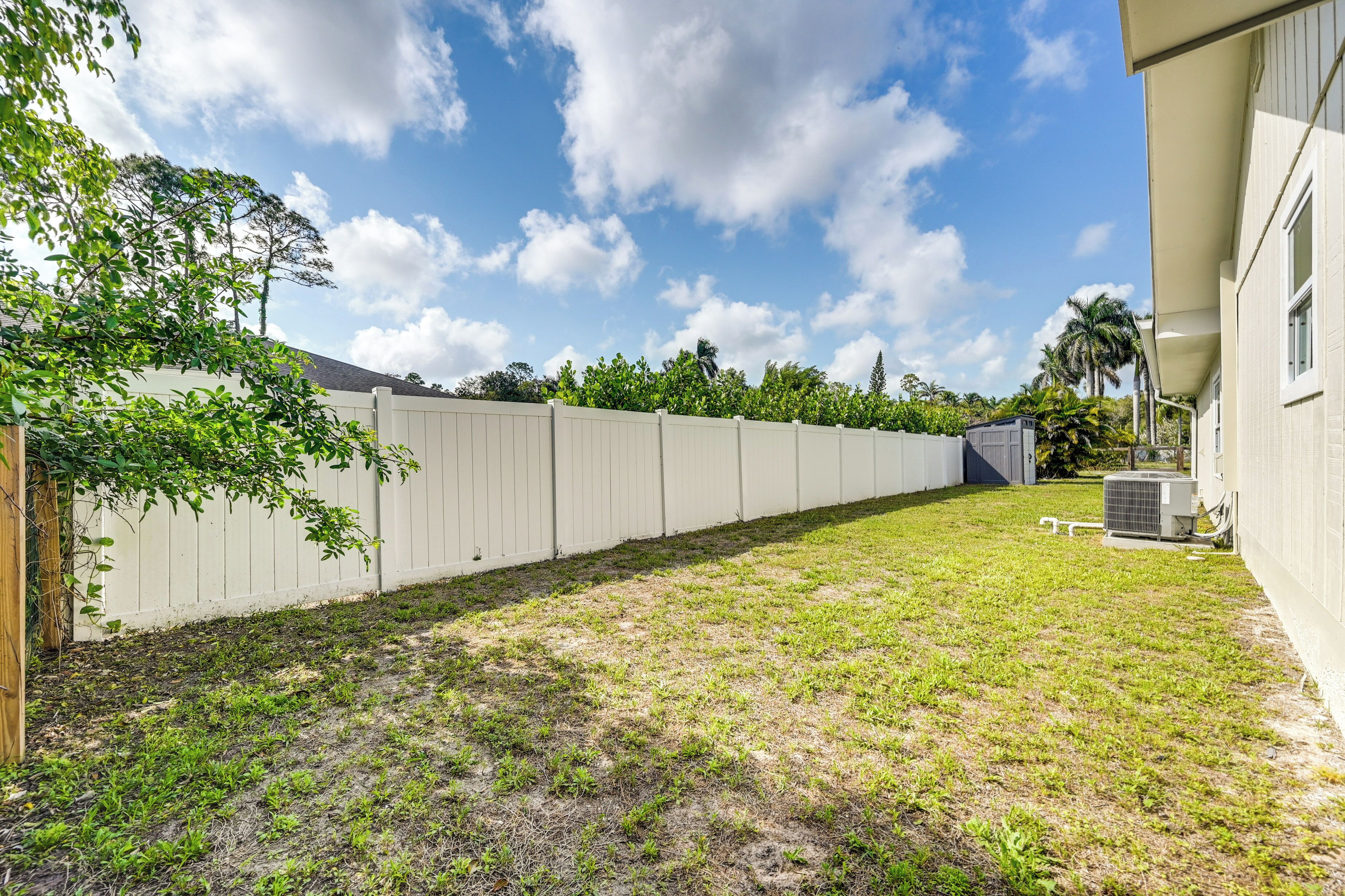 A fenced backyard with a grassy area, a white building, and an air conditioning unit.