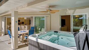 A hot tub with a ceiling fan, a dining area with chairs and a table, and a kitchen visible through glass doors.