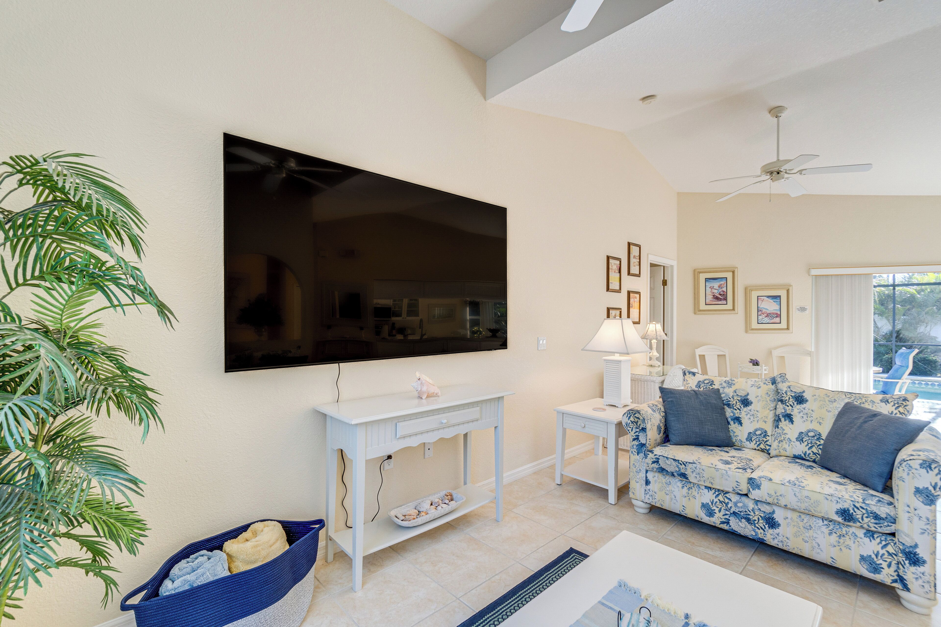 Un salon avec un téléviseur à écran plat, une console blanche, un canapé à motifs floraux, un panier bleu en osier et une plante en pot.