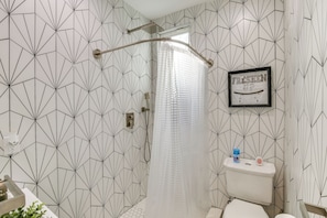 Um banheiro com parede estampada em padrão geométrico, chuveiro com cortina transparente, vaso sanitário e uma placa emoldurada na parede.
