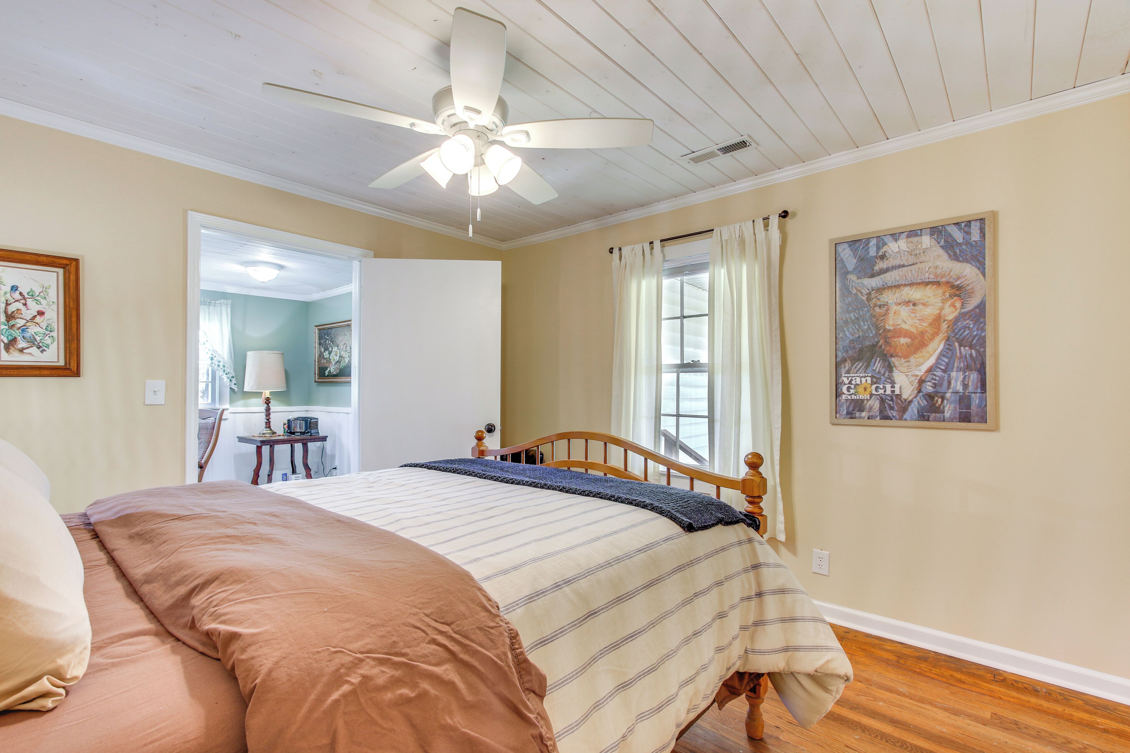 A bedroom with a bed, a ceiling fan, a window with curtains, and a painting on the wall.