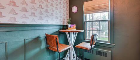 A corner room with two chairs, a small table, a clock, and a window with blinds.