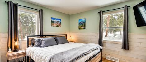 Un dormitorio con cama, mesita de noche, lámpara y dos ventanas con cortinas.