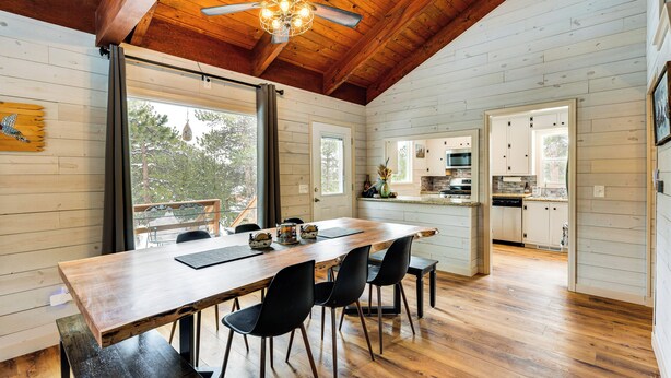 A spacious dining area with a wooden table and chairs, a ceiling fan, and large windows offering a view of the outdoors. - Quiet + Peaceful Home in Allenspark w/ Deck! (Allenspark)