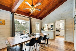 Un comedor amplio con una mesa y sillas de madera, un ventilador de techo y ventanas grandes que ofrecen vistas al exterior.