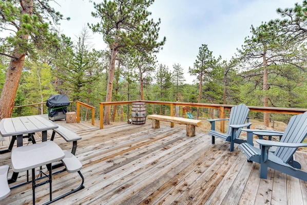 Una terraza de madera con mesas de picnic y sillones Adirondack, rodeada de árboles.