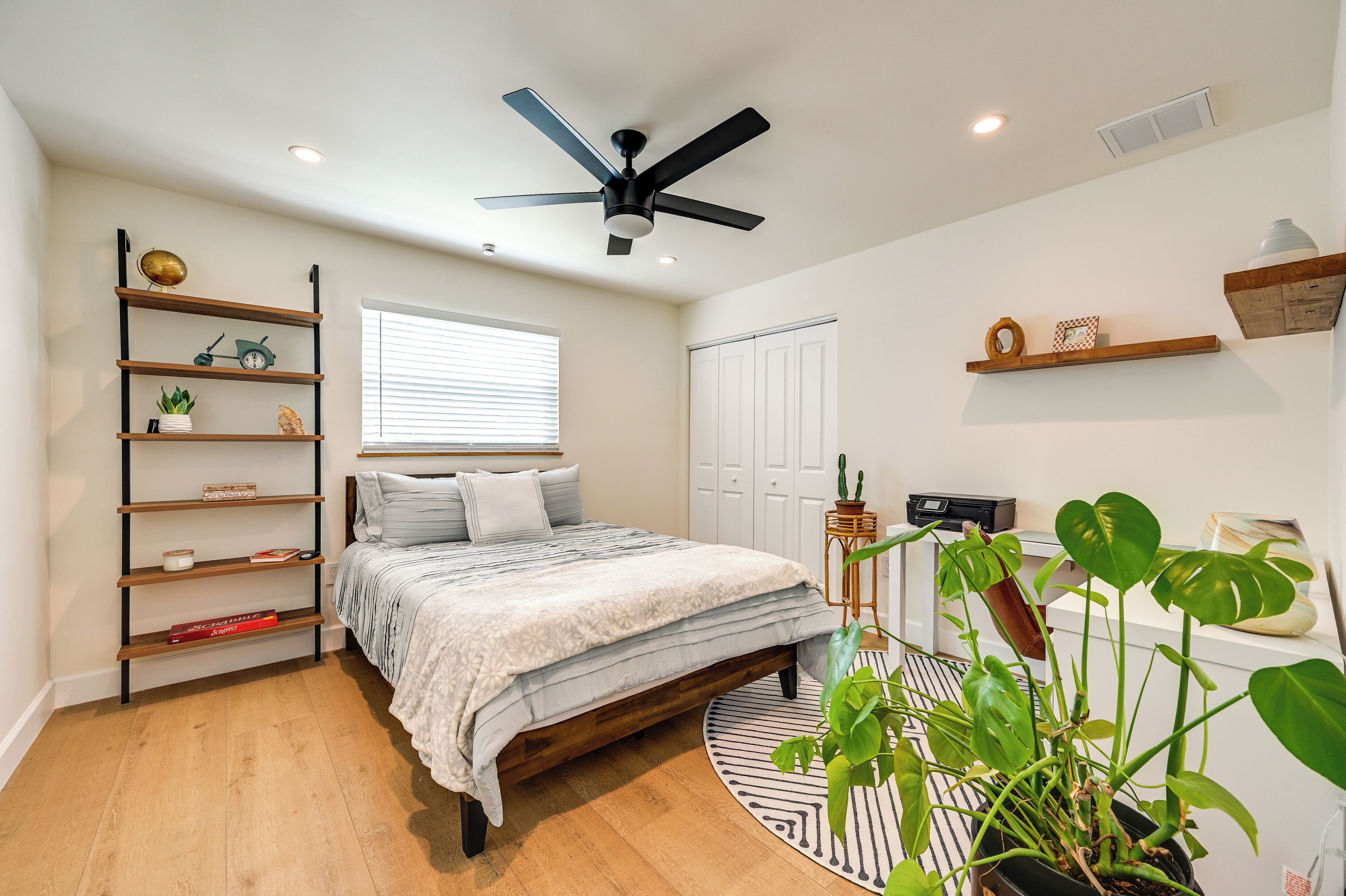 A bedroom with a bed, a ceiling fan, a window, a wardrobe, shelves with plants, and a potted plant.