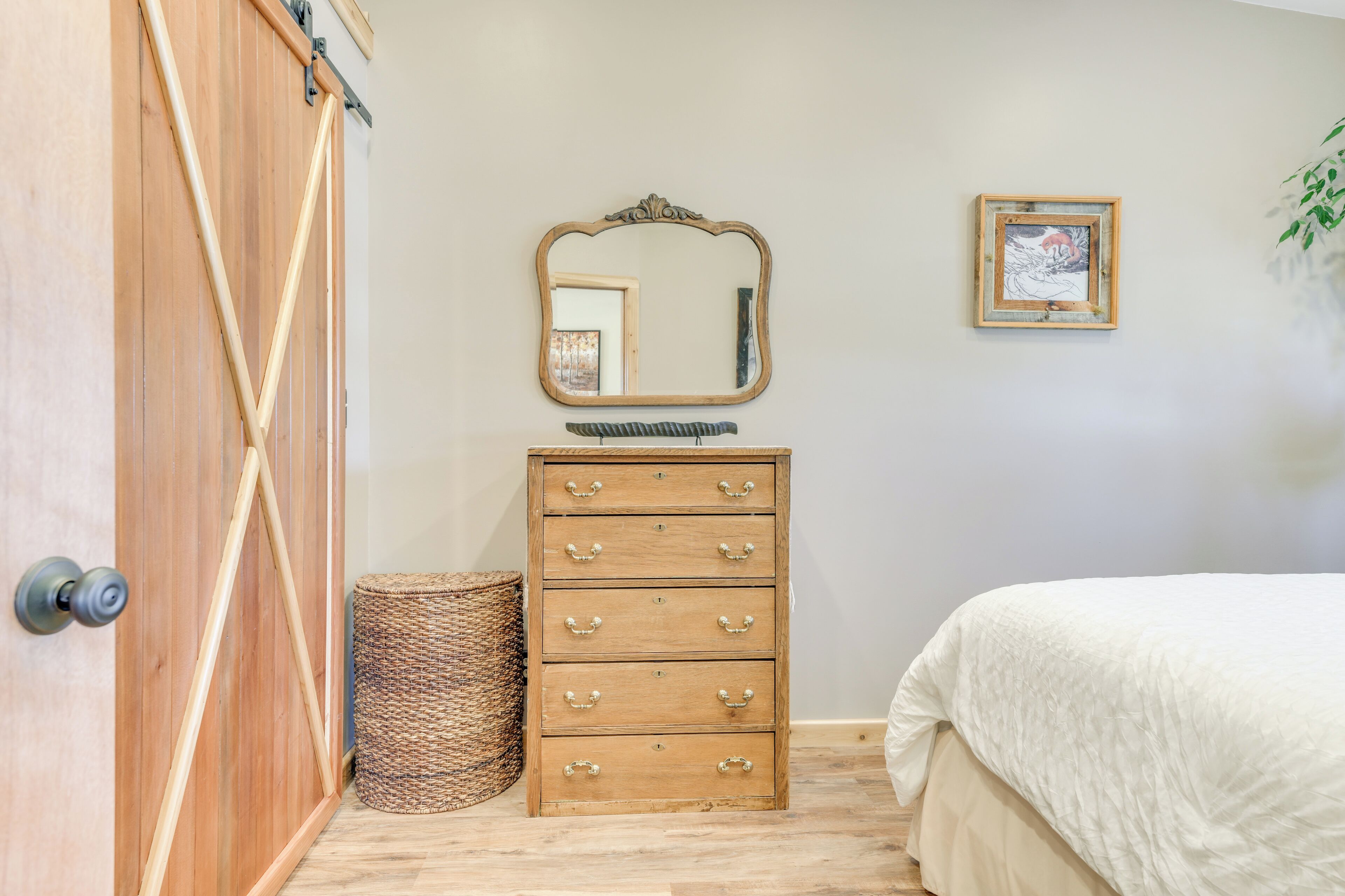 A bedroom with a wooden dresser, a mirror, a framed picture, a wicker basket, and a bed with a white quilt.