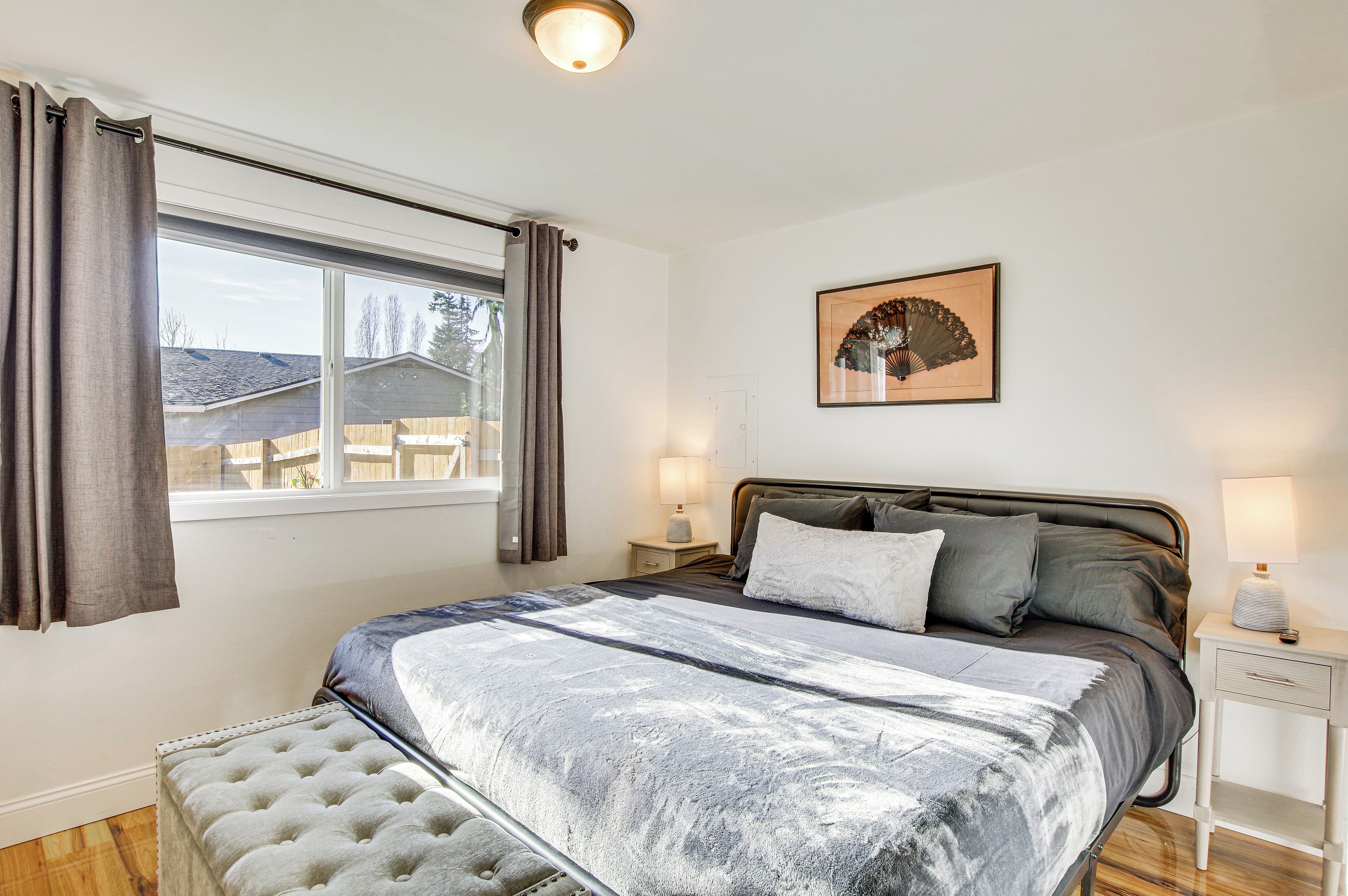 Un dormitorio con cama, mesitas de noche, ventana con cortinas, un ventilador de pared y un cuadro enmarcado.