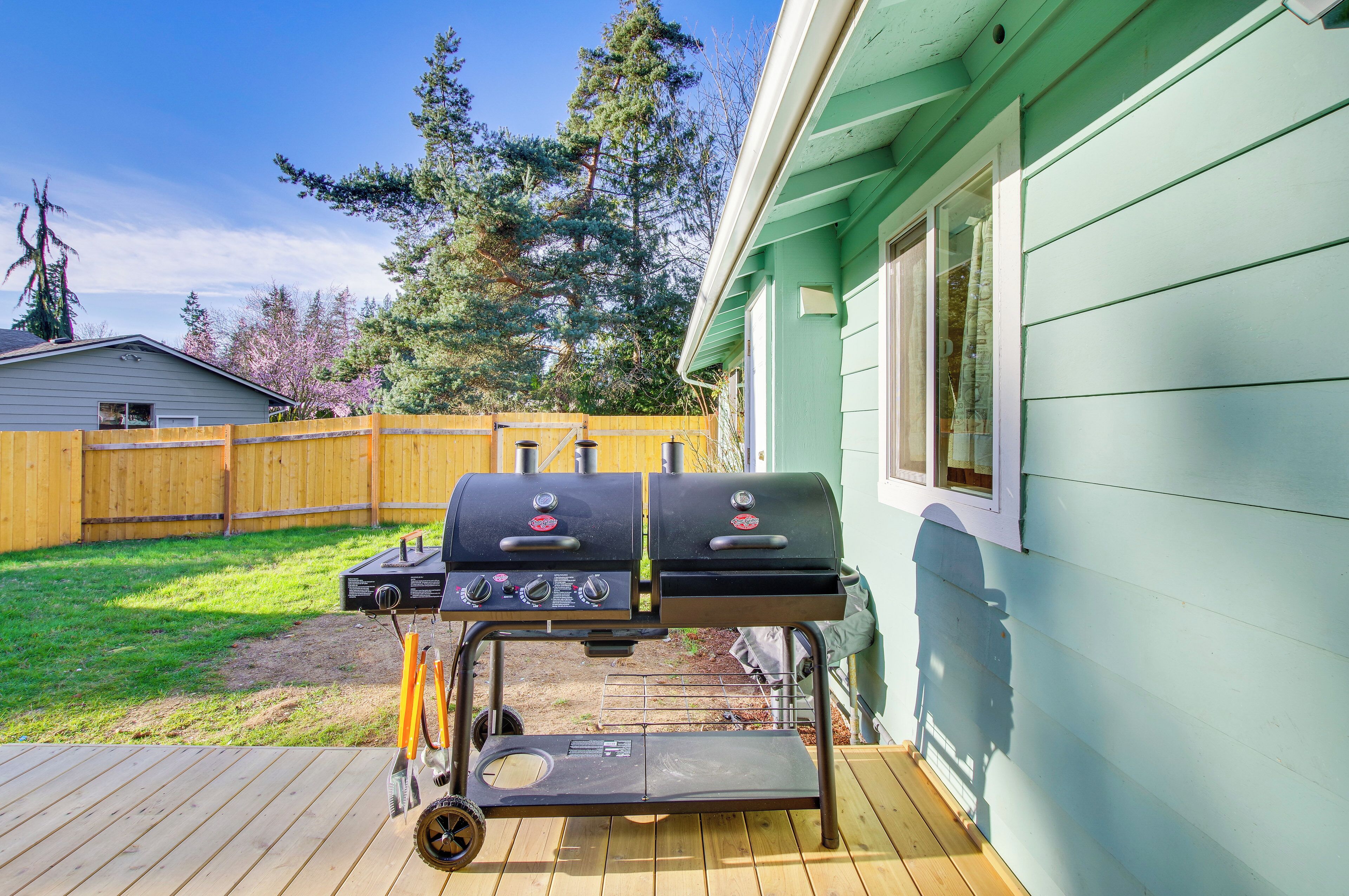 Ein Garten mit einem Grill mit zwei Ebenen, einer Holzterrasse und einem Gewächshaus.