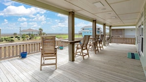 A covered outdoor area with wooden chairs and tables, a wooden deck, and a view of the beach and houses in the distance.