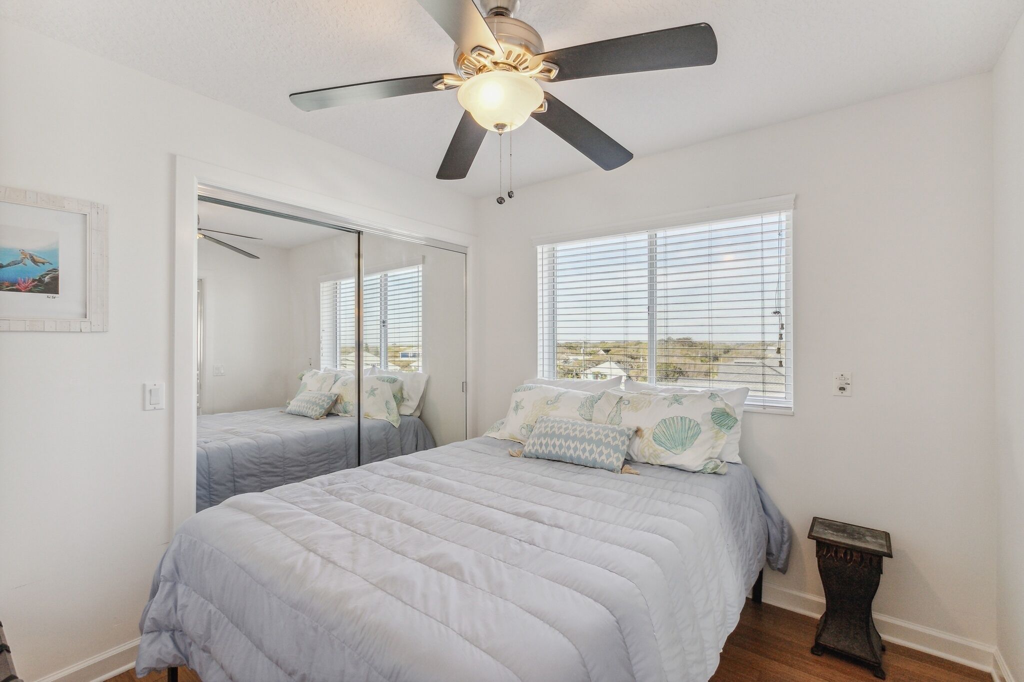 Une chambre à coucher avec un lit, un ventilateur de plafond, une fenêtre avec des stores et une petite table.