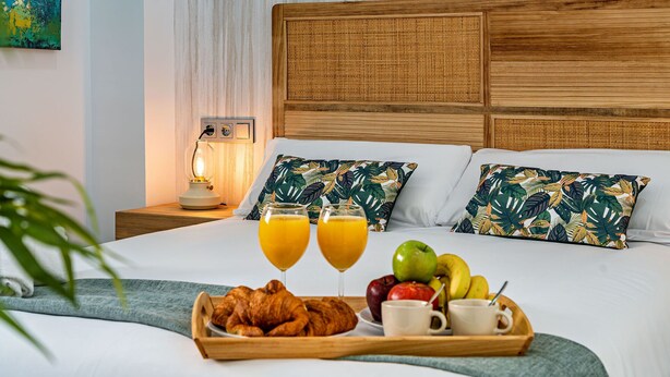 Ein ordentlich bezogenes Bett mit Holz-Kopfstück, zwei Kissen mit tropischem Blattmuster, ein Nachttisch mit Lampe und eine Ablage mit Croissants, Obst und Orangensaft.